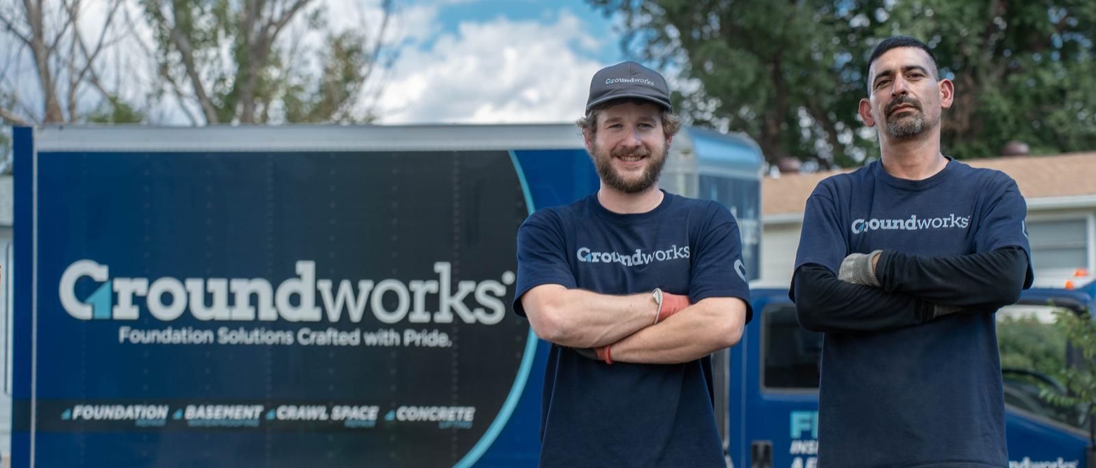 Two men in Groundworks shirts stand in front of a Groundworks truck on a sunny day, smiling at the camera.