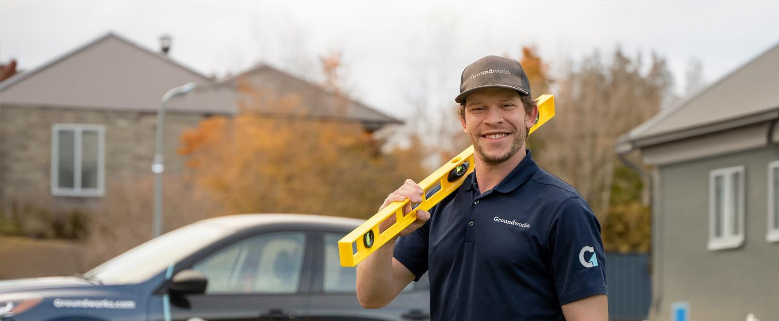 A smiling man in a Groundworks uniform holds a yellow level tool, standing in front of a Groundworks vehicle.
