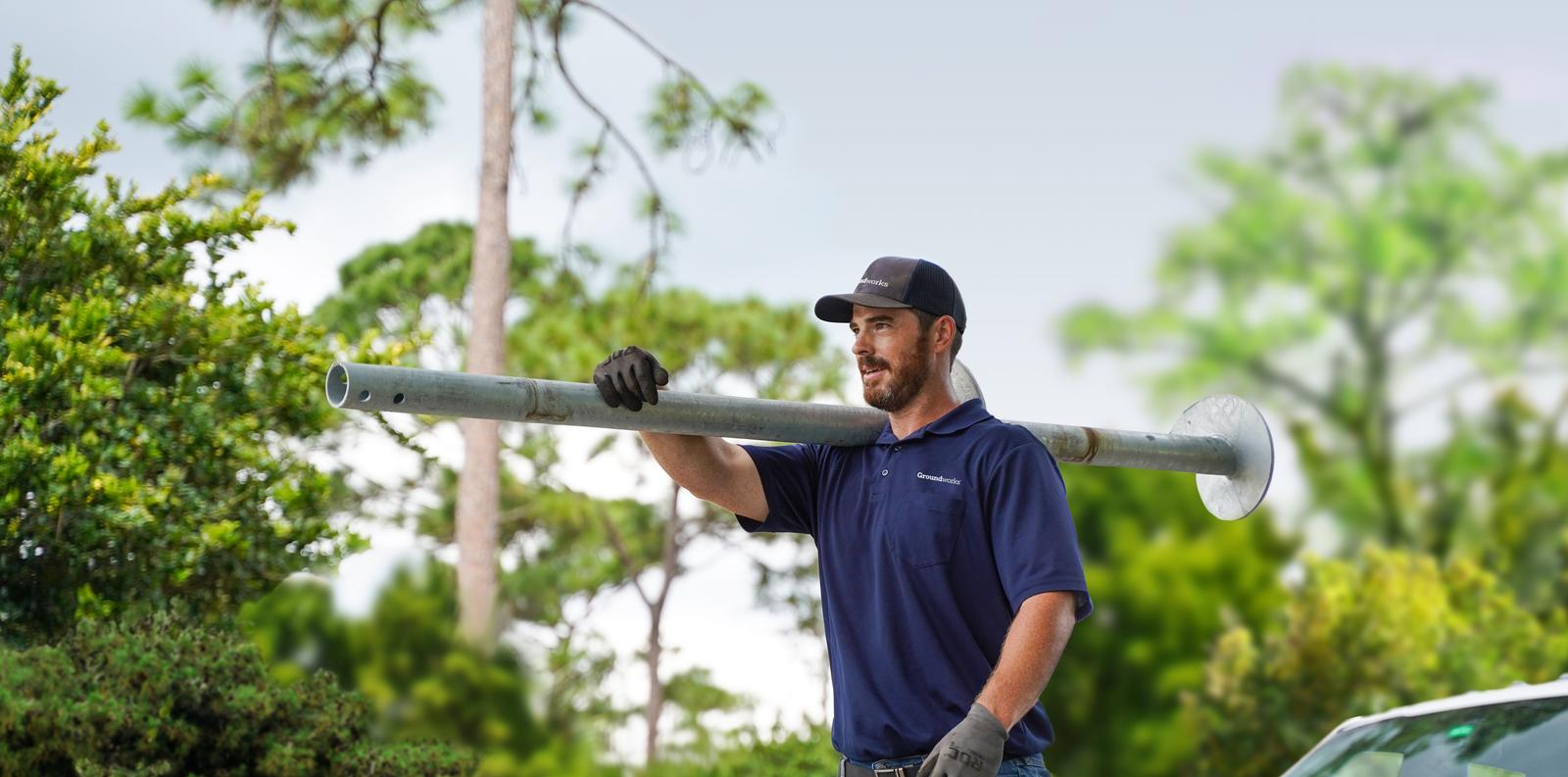 A man in work gloves and a cap carries a large metal pole on his shoulder outdoors, with trees and a white truck nearby.