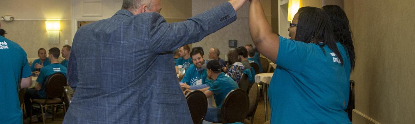 Two people in teal shirts high-five in a room with seated attendees.