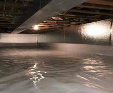 The crawl space, with reflective insulation and a vapor barrier on the floor, is softly illuminated by a single light bulb. Strategically placed crawl space vents ensure proper airflow and maintain an optimal environment.