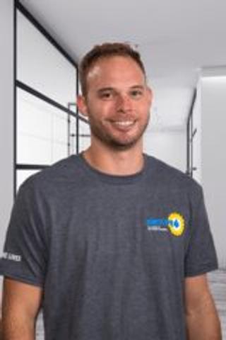 Smiling man in a grey t-shirt stands in a modern hallway with glass partitions.