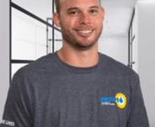 Smiling man in a grey t-shirt stands in a modern hallway with glass partitions.