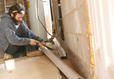 Person installing a gutter system on a basement floor near a brick wall.