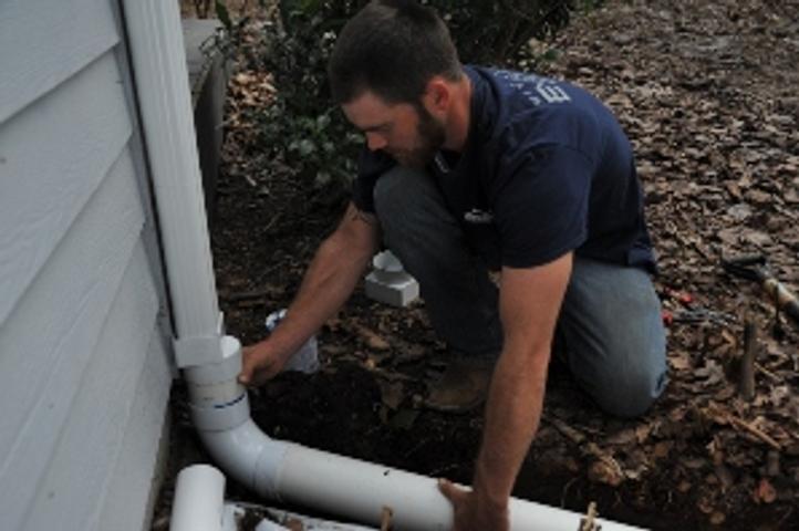 Person fixing outdoor pipe near house wall in a garden area.