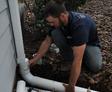 Person fixing outdoor pipe near house wall in a garden area.
