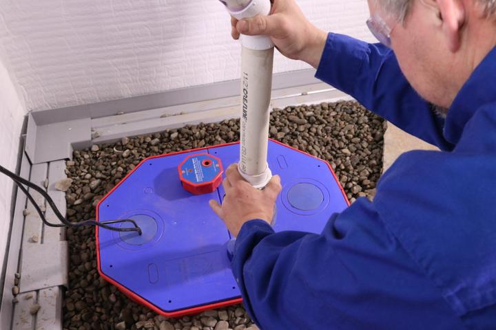 Person installing a white PVC pipe into a blue sump pump cover surrounded by gravel.