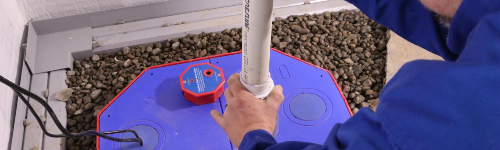 Person installing a white PVC pipe into a blue sump pump cover surrounded by gravel.