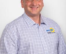 Smiling person in a checkered shirt stands against a plain background.