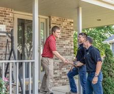 Three men shake hands on a porch, engaged in conversation near a brick house entrance.