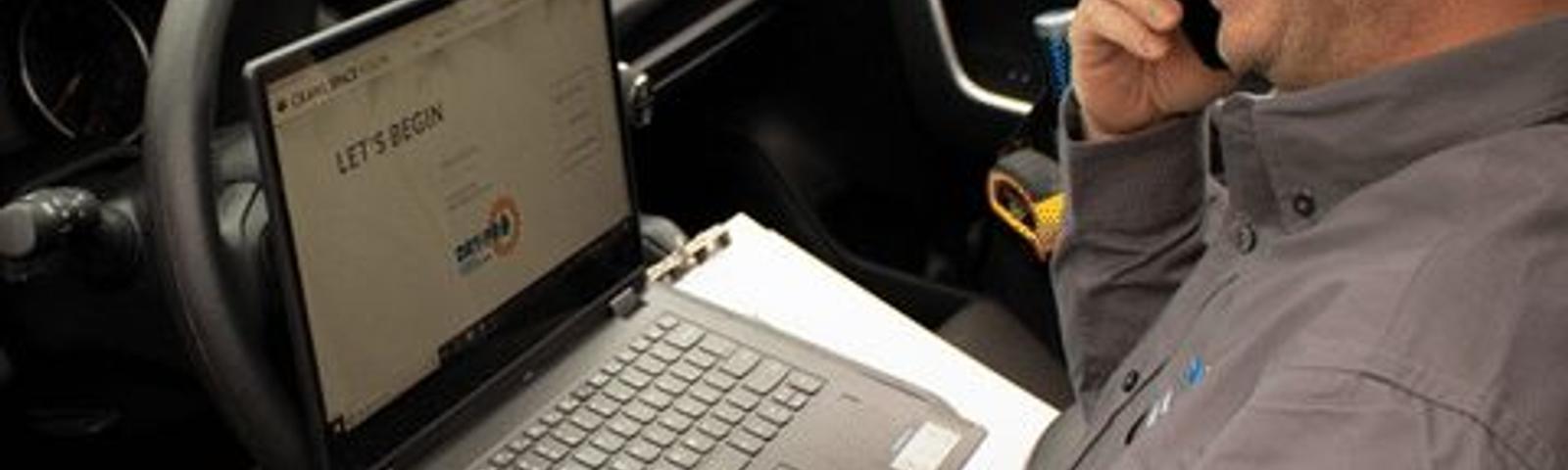 Man in a car taking notes on a laptop, smiling while on a phone call. "DRY-PRO" logo visible on screen and image.