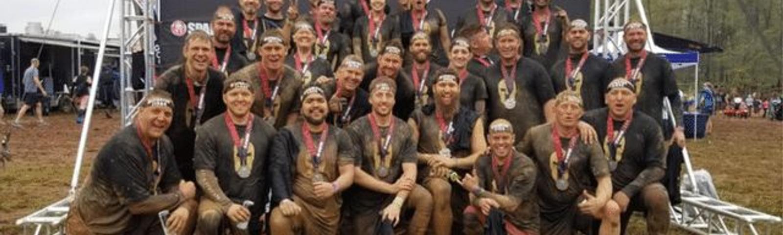Group of muddy athletes with medals pose under a Spartan Race banner.