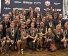 Group of muddy athletes with medals pose under a Spartan Race banner.