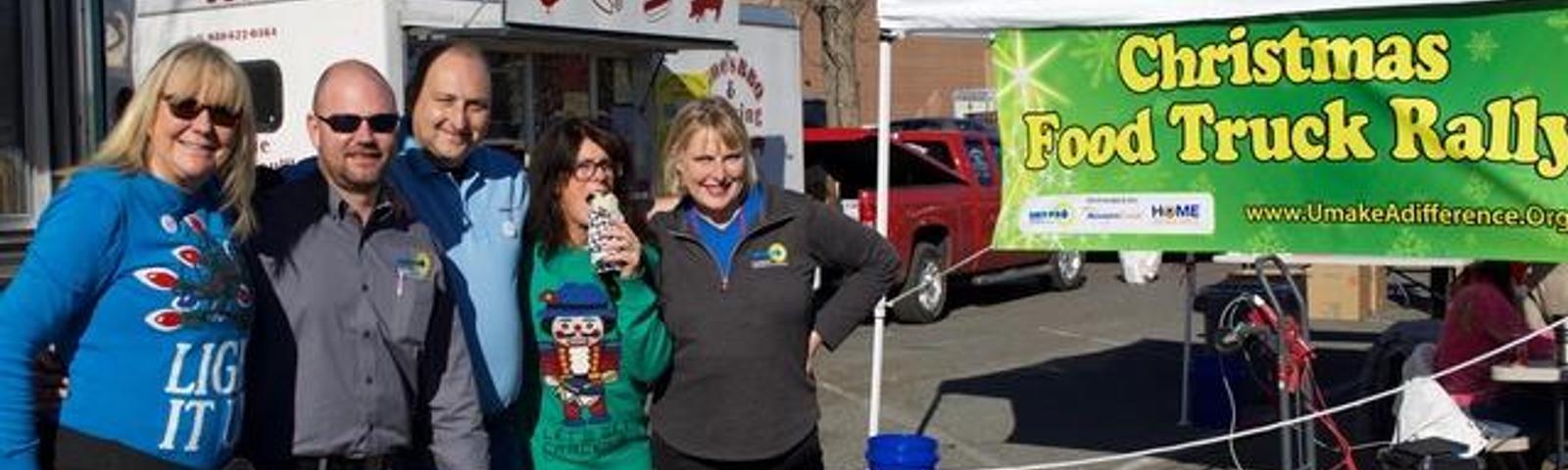 A group of people at a Christmas Food Truck Rally under a sunny sky, with food trucks and a banner in the background.