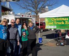 A group of people at a Christmas Food Truck Rally under a sunny sky, with food trucks and a banner in the background.