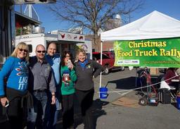 A group of people at a Christmas Food Truck Rally under a sunny sky, with food trucks and a banner in the background.
