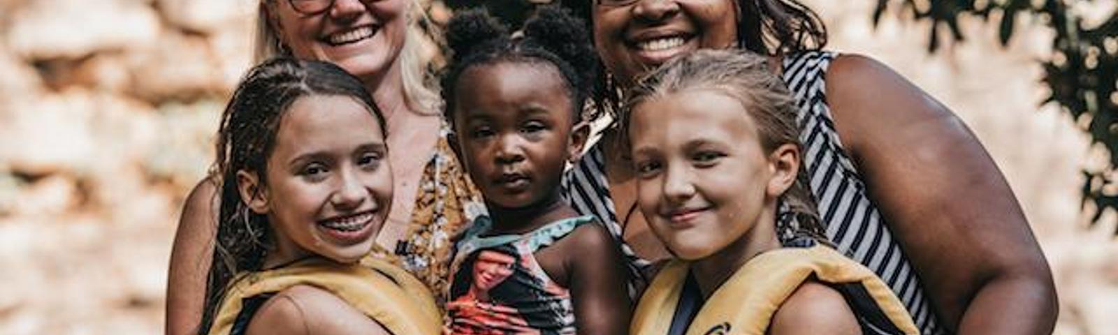 Two women and three children in life jackets smile outdoors near water and trees.