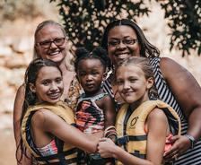 Two women and three children in life jackets smile outdoors near water and trees.