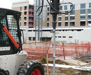 Construction site with a drilling machine creating a hole in the ground, surrounded by buildings and safety barriers.