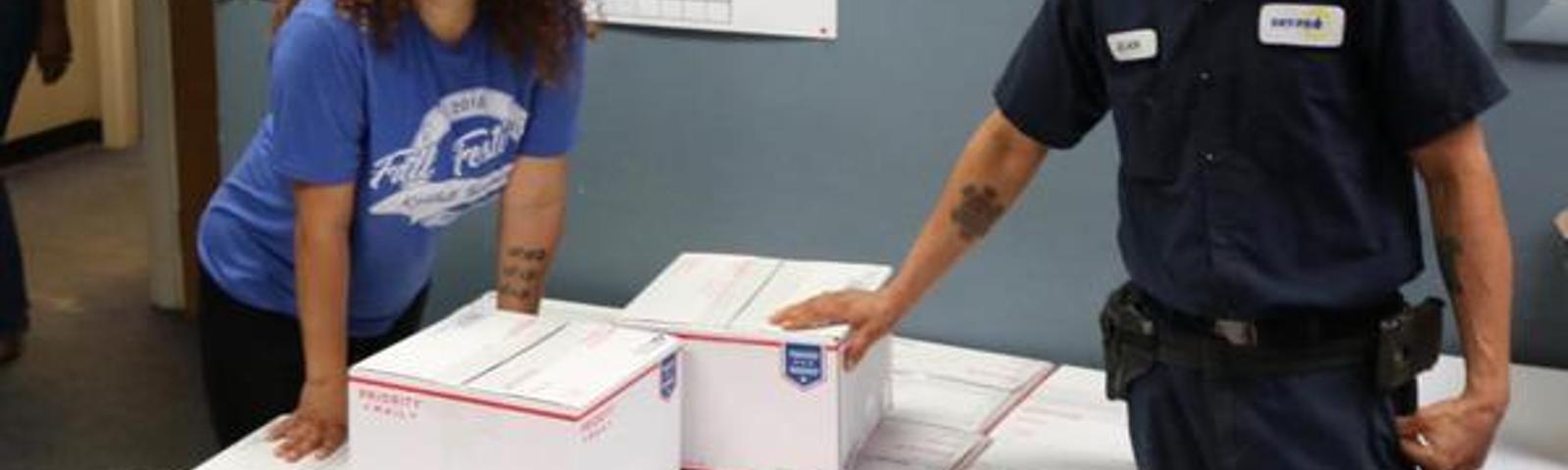 Two people standing by a stack of white boxes in a room with a wall chart.