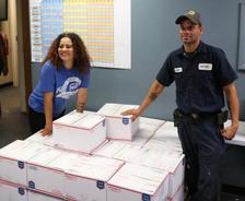 Two people standing by a stack of white boxes in a room with a wall chart.