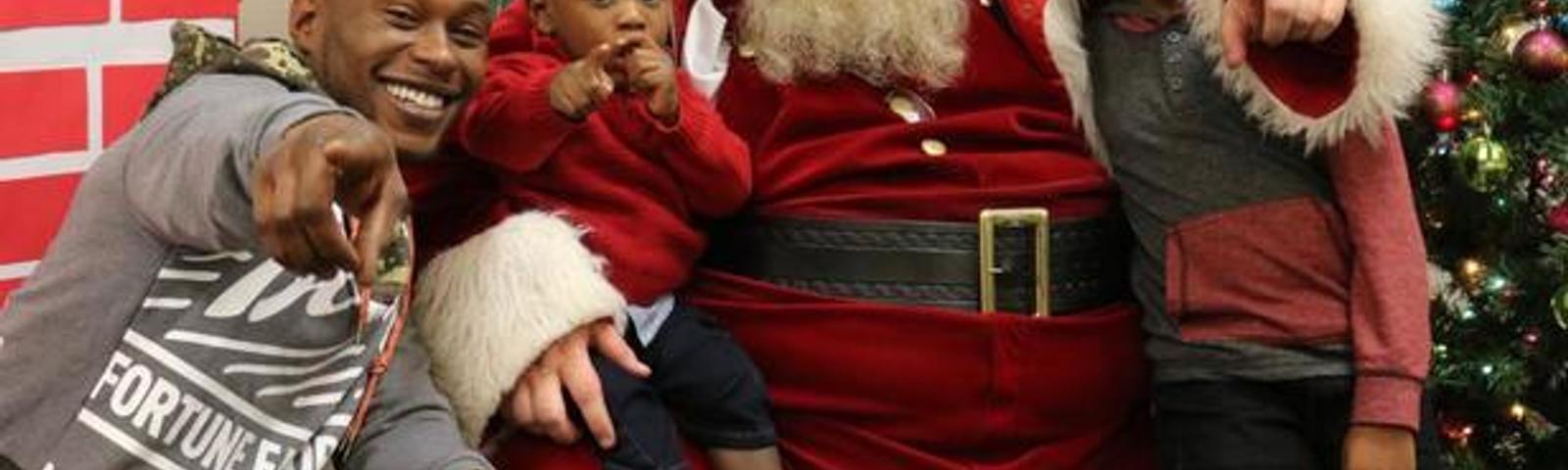 A man and two children pose with Santa Claus, all smiling and pointing, near a Christmas tree indoors.