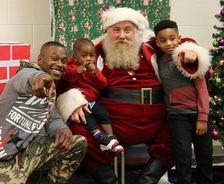 A man and two children pose with Santa Claus, all smiling and pointing, near a Christmas tree indoors.