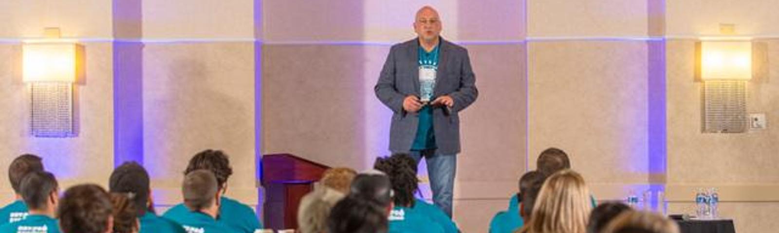 A speaker presents to an audience wearing matching teal shirts in a conference room.