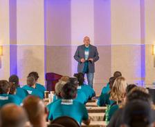 A speaker presents to an audience wearing matching teal shirts in a conference room.