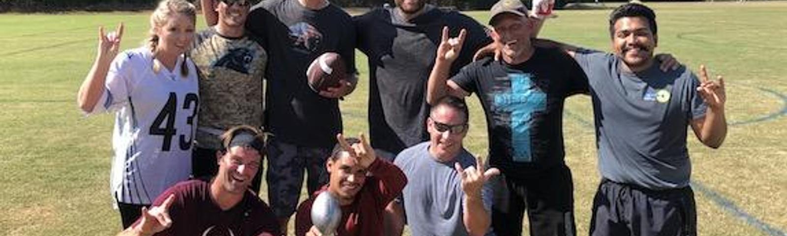 A group of people outdoors smiling, some holding a football, with trees and a field in the background.
