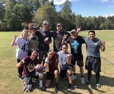 A group of people outdoors smiling, some holding a football, with trees and a field in the background.