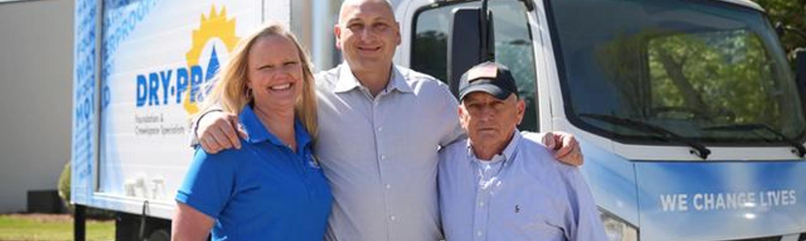 Three people stand smiling in front of a Dry Pro truck with the slogan "We Change Lives" on it.