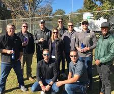 Group of people posing outdoors in casual clothing, some holding drinks, with a fence and trees in the background.
