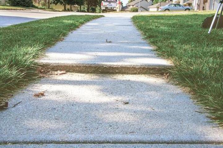 A concrete sidewalk with an uneven surface surrounded by green grass and a street in the background.