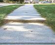 A concrete sidewalk with an uneven surface surrounded by green grass and a street in the background.