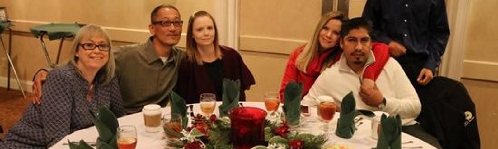 Group of six people seated around a decorated round table with drinks and holiday centerpieces.