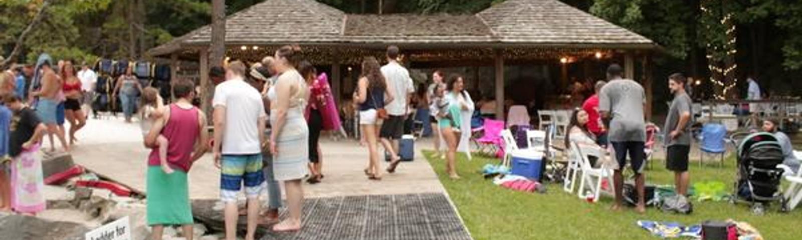 People gather near a lakeside pavilion, surrounded by trees, chairs, and pool noodles on a sunny day.