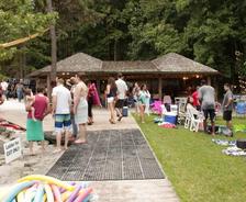 People gather near a lakeside pavilion, surrounded by trees, chairs, and pool noodles on a sunny day.