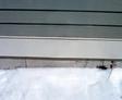 A snow-covered ground next to a gray wall with horizontal lines and a single small plant stem visible.