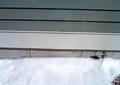 A snow-covered ground next to a gray wall with horizontal lines and a single small plant stem visible.