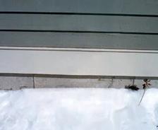 A snow-covered ground next to a gray wall with horizontal lines and a single small plant stem visible.