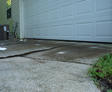Cracked concrete driveway in front of a white garage door at a house, with grass on the side.