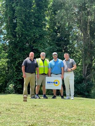 Four men standing on grass with a DRY-PRO sign, surrounded by trees.