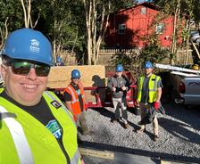 Four construction workers in safety vests and helmets pose on a gravel driveway with a trailer in the background.