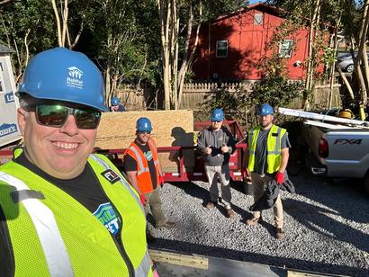 Four construction workers in safety vests and helmets pose on a gravel driveway with a trailer in the background.