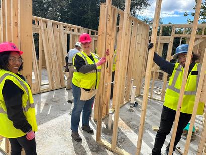 Three people in hard hats and vests work on a wooden house frame at a construction site.