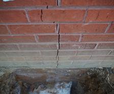Vertical crack in a red brick wall with exposed ground at the base of the wall.