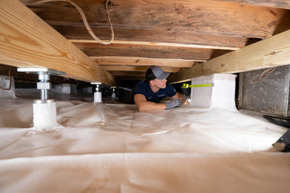 Person measuring space under wooden floor beams, surrounded by white insulation material.