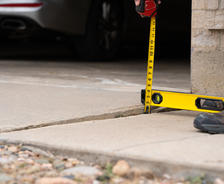 Person measuring uneven sidewalk with a tape measure and yellow level tool near a brick wall.