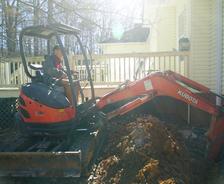 A person uses an orange Kubota mini excavator for basement waterproofing next to a yellow house and wooden deck.